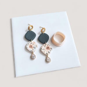 May include: Dangle earrings with a floral design and crystal teardrop details. The earrings have dark teal and floral patterned sections, with gold-tone metal components. A light pink, circular clay cutter is also visible.