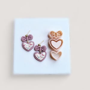 May include: A pair of mauve heart-shaped earrings with floral patterns and bow accents, alongside heart-shaped cookie cutters. The earrings have gold-colored posts. The cookie cutters are in the shape of a bow and hearts.