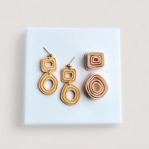 May include: Gold-coloured, geometric dangle earrings with a square top and a circular bottom. Two additional clay shapes, a square and a circular design, are also present. The earrings and shapes are displayed on a white surface.