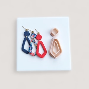May include: Three pairs of handmade earrings in a variety of colors and patterns, including navy blue, floral, and red. The earrings are displayed on a white surface with two matching clay cutters.