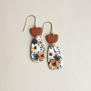 May include: A pair of dangle earrings featuring a textured, semi-circular terracotta-colored top piece connected to a teardrop-shaped pendant. The pendant displays a floral pattern with black, white, and orange flowers on a light background.