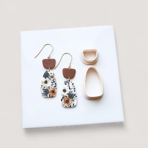 May include: Handmade earrings with a floral pattern and gold-coloured hooks. The earrings have a brown textured top and a white, black, and brown floral design. Two earring-shaped clay cutters are also visible.