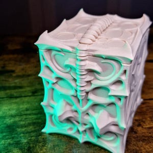 May include: A detailed, off-white decorative box with an intricate, gothic-inspired design. The box features a series of layered, curved patterns and a central spine-like detail on the lid. The box is illuminated with a green light, creating a dramatic effect.