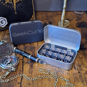 Könnte beinhalten: Ein Set aus zehn dunkelblau marmorierten Würfeln mit goldfarbenen Zahlenmarkierungen, untergebracht in einer rechteckigen silbernen Metalldose mit dem aufgedruckten Text GeekCurio auf dem Deckel. Die Würfel zeigen verschiedene Symbole.