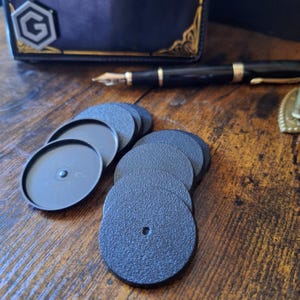 May include: A collection of small, circular black plastic bases with a textured top surface and a central hole. These round components feature a raised rim and are designed for mounting miniature figures or similar small objects for display or gaming purposes.