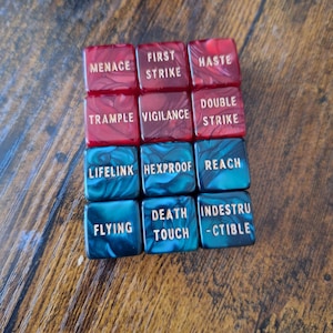 May include: A set of twelve square game tokens arranged in a grid, featuring six red and six blue marbled acrylic pieces. Each token is engraved with gold text including Menace, First Strike, Haste, Trample, Vigilance, Double Strike, Lifelink, Hexproof, Reach, Flying, Deathtouch, and Indestructible.