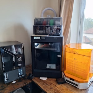May include: A selection of 3D printing machines. The image shows a black printer with a clear door, a black printer with the number "05" on the front, and an orange resin curing machine. The printers are on a wooden surface.