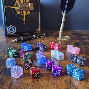 May include: A collection of multi-coloured six-sided dice featuring various numerical modifiers printed in white. The dice are scattered across a surface in front of a black journal with a gold compass design, a fountain pen, and a quill pen stand.