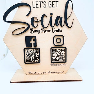 Può includere: Segno esagonale in legno con il testo "Let's Get Social" e le parole "Busy Bear Crafts" sotto. Il segno ha due codici QR, uno per Facebook e uno per Instagram, con le icone dei social media sopra ogni codice. Il segno ha anche il testo "@busybearcrafts" e "Grazie per essere passato!"