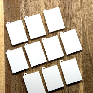 May include: A collection of nine rectangular white acrylic pendants, each featuring a small circular loop at the top edge for attachment. These blank, flat, and smooth-surfaced components are arranged in a grid-like pattern, showcasing a uniform design.