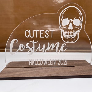 May include: A clear acrylic Halloween decoration with the words "Cutest Costume" and a skull design. The base is made of dark wood, and the text "Halloween 2021" is also included.