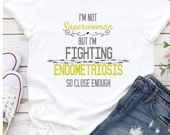 May include: White t-shirt with the text "I'm not Superwoman but I'm fighting Endometriosis so close enough". The text is in yellow and gray. The shirt is on a white wooden surface with a pair of white sneakers and blue denim shorts.