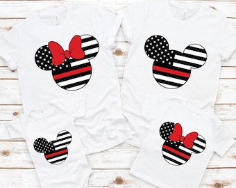 Könnte beinhalten: Vier weiße T-Shirts mit einem rot-weiß-blauen Mickey-Maus-Motiv im Stil der amerikanischen Flagge. Das Design enthält eine dünne rote Linie in der Mitte der Flagge, die Feuerwehrleute repräsentiert. Zwei der Shirts haben eine rote Schleife auf dem Mickey-Maus-Kopf, während die anderen beiden keine haben.