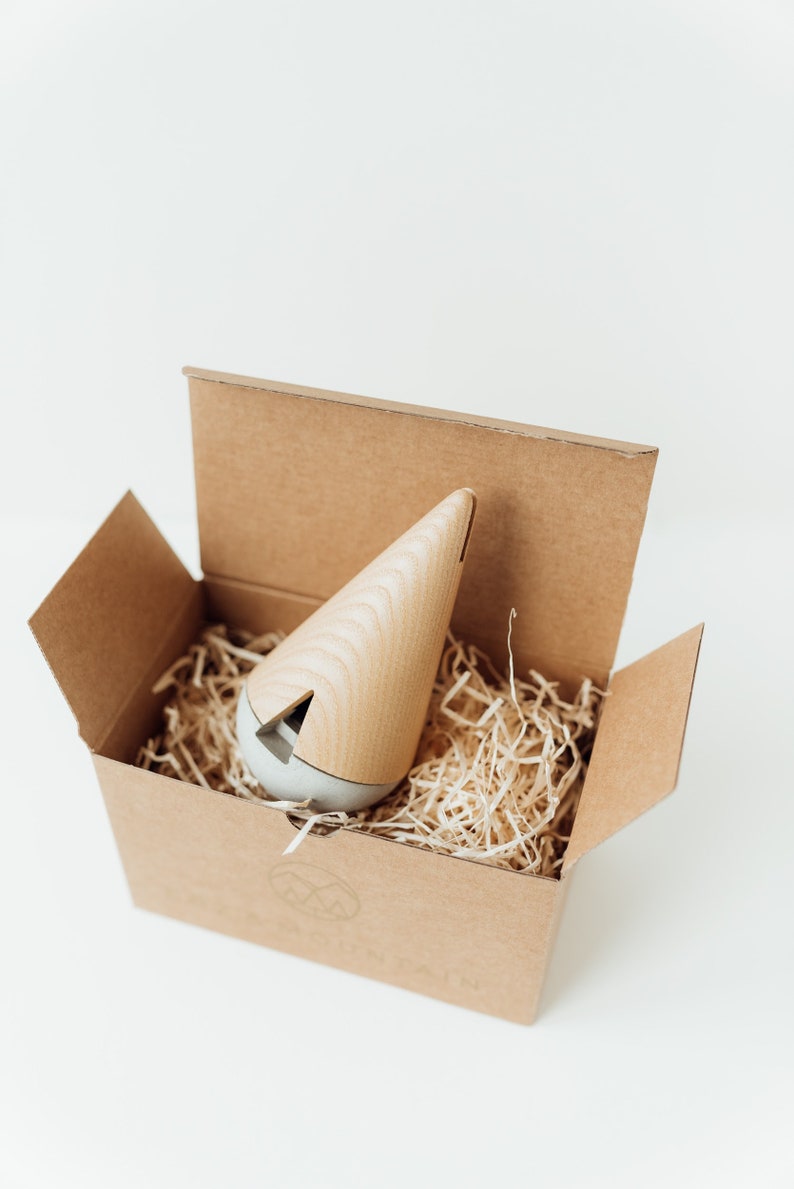 May include: A wooden and gray cone-shaped incense burner sits in a brown cardboard box filled with shredded paper. The box has a logo with two mountains and the word "Mountain" printed on it.