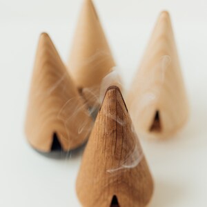 May include: Four wooden cone incense burners with smoke rising from the top of one. The cones are a light brown color and have a small opening at the bottom.