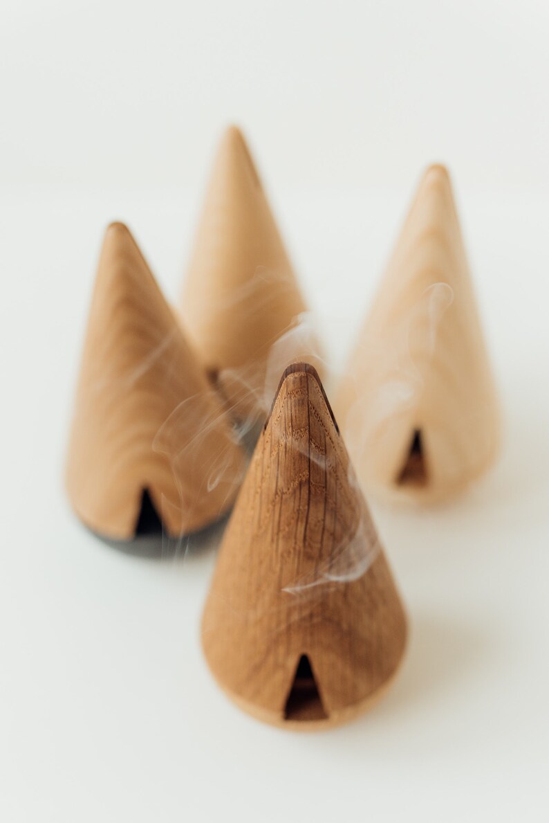 May include: Four wooden cone incense burners with smoke rising from the top of one. The cones are a light brown color and have a small opening at the bottom.