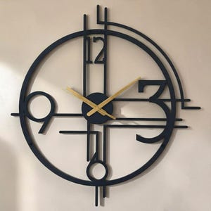 Könnte beinhalten: Eine runde Wanduhr mit einem schwarzen Metallrahmen, geometrischen Linien und offenen Bereichen. Das Zifferblatt zeigt die Zahlen 12, 3, 6 und 9 in einer fetten Schrift. Zwei dünne, goldfarbene Zeiger befinden sich in der Mitte der Uhr.