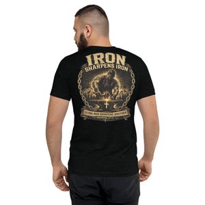 May include: A black short-sleeve t-shirt featuring a graphic on the back. The design shows a blacksmith at an anvil with the text IRON SHARPENS IRON and SO ONE MAN SHARPENS ANOTHER PROVERBS 27:17, framed by a chain and a cross symbol.