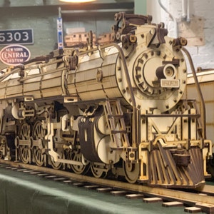 Puede incluir: Un modelo de madera detallado de una locomotora de vapor, pintada de blanco con detalles negros. El modelo presenta detalles intrincados, incluyendo ruedas, engranajes y una chimenea. La locomotora está etiquetada con "5303" y "New York Central System".