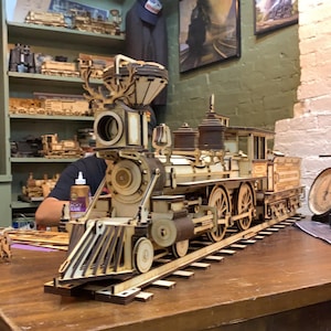 May include: A detailed wooden model of a vintage steam locomotive train engine. The model is assembled and sitting on a wooden table. The train engine is made of wood and has a brown and cream color scheme.