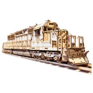 May include: A detailed wooden model of a diesel locomotive, featuring intricate cut-out designs. The model is light brown and sits on a section of model train tracks. The locomotive has the text "RDX 2025" on the side.