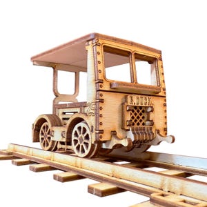 May include: A wooden model train with intricate details, including a roof, windows, and wheels. The model is on a wooden track, with the number "25" visible on the side and the text "R. DUCK" on the back. The train is made of light brown wood.
