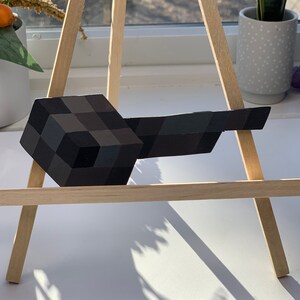 May include: A black and gray Minecraft pickaxe made of wood and painted to look like a pixelated block. The pickaxe is resting on a wooden easel.
