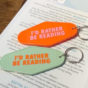 May include: Two retro key fobs with the text "I'D RATHER BE READING" in orange. One is orange, the other mint green. Both have silver key rings. They rest on an open book. The key fobs are a fun accessory.