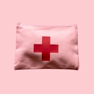 Puede incluir: Una bolsa con cremallera de lona rosa claro con una gran cruz roja en el centro. La bolsa es rectangular y parece estar hecha de una tela texturizada. El fondo es de un rosa claro a juego.