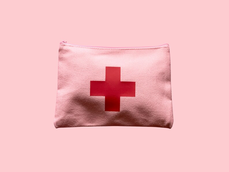 Puede incluir: Una bolsa con cremallera de lona rosa claro con una gran cruz roja en el centro. La bolsa es rectangular y parece estar hecha de una tela texturizada. El fondo es de un rosa claro a juego.