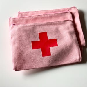 Puede incluir: Tres estuches con cremallera de lona rosa claro, cada uno con una cruz roja en la parte delantera. Los estuches est&aacute;n apilados, con el superior solapando ligeramente a los dem&aacute;s. Las cremalleras son rosas.
