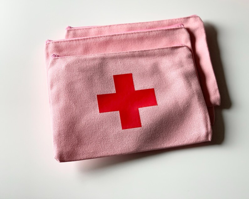 Puede incluir: Tres estuches con cremallera de lona rosa claro, cada uno con una cruz roja en la parte delantera. Los estuches est&aacute;n apilados, con el superior solapando ligeramente a los dem&aacute;s. Las cremalleras son rosas.