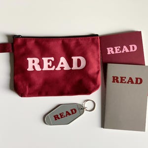 Peut inclure: Ensemble d'articles sur le thème de la lecture : une pochette zippée bordeaux, deux carnets et un porte-clés, tous avec le mot "READ". La pochette et le porte-clés ont une police rose clair, tandis que les carnets ont une police rouge plus foncée. Sur fond blanc.