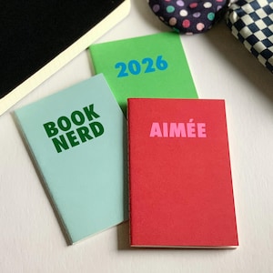 May include: Three small rectangular notebooks with paper covers. One is light blue with green text reading BOOK NERD, one is bright green with blue text reading 2026, and one is red with pink text reading AIMÉE. The notebooks are arranged together.