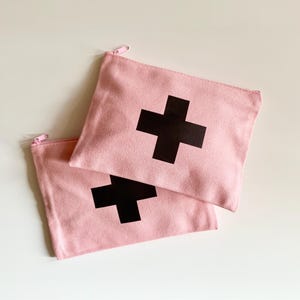 Puede incluir: Dos estuches con cremallera de lona rosa con una gran cruz negra estampada en la parte delantera. Los estuches son rectangulares y tienen una cremallera rosa claro. El fondo es una superficie blanca lisa.
