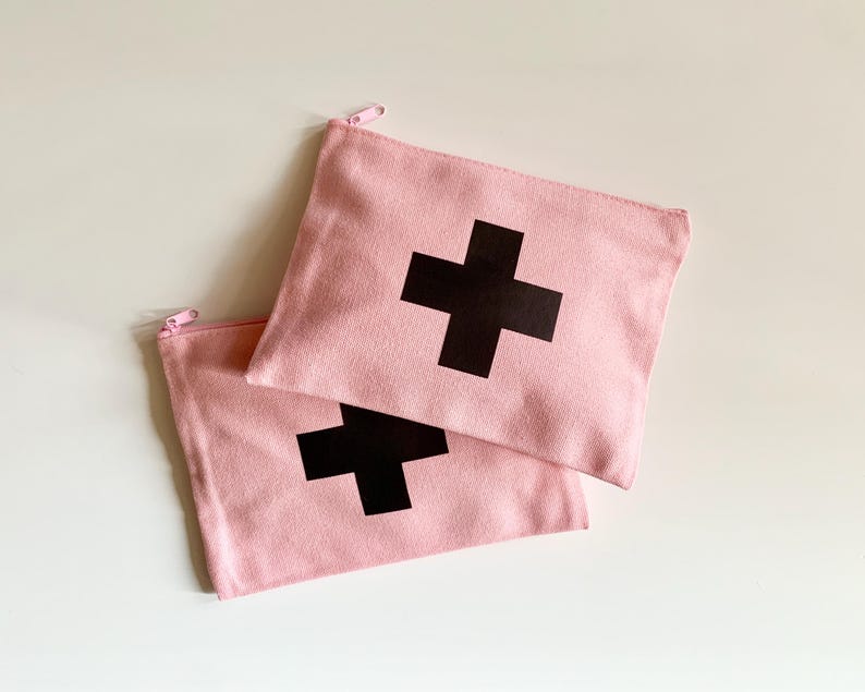 Puede incluir: Dos estuches con cremallera de lona rosa con una gran cruz negra estampada en la parte delantera. Los estuches son rectangulares y tienen una cremallera rosa claro. El fondo es una superficie blanca lisa.