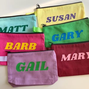 Peut inclure: Plusieurs pochettes zippées en toile colorées, chacune affichant un nom différent en lettres contrastées et audacieuses. Les pochettes sont disponibles en teal, rose, jaune, vert, bordeaux et lavande. Les noms sont "Matt", "Barb", "Susan", "Gary", "Mary" et "Gail".