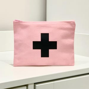 Puede incluir: Bolsa de lona rosa con un dise&ntilde;o de cruz negra. La bolsa rectangular tiene cierre de cremallera. La bolsa est&aacute; sobre una superficie blanca.