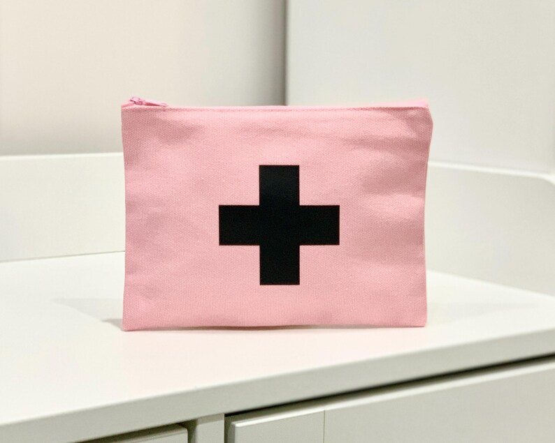 Puede incluir: Bolsa de lona rosa con un dise&ntilde;o de cruz negra. La bolsa rectangular tiene cierre de cremallera. La bolsa est&aacute; sobre una superficie blanca.