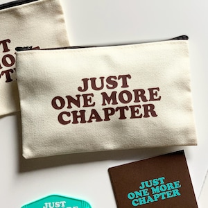 Peut inclure: Pochette zippée en toile écrue avec l'inscription "JUST ONE MORE CHAPTER" en marron. Une deuxième pochette et plusieurs cartes avec le même texte sont également visibles. Les pochettes ont des fermetures éclair noires.