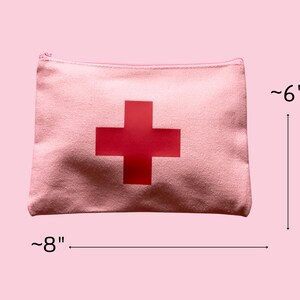 Puede incluir: Una bolsa con cremallera de lona rosa con una gran cruz roja en el centro. La bolsa mide aproximadamente 20 cm de ancho y 15 cm de alto. La bolsa de tem&aacute;tica m&eacute;dica est&aacute; dise&ntilde;ada para guardar suministros de primeros auxilios.
