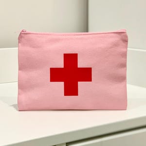 Puede incluir: Una bolsa de lona rosa con cremallera y una gran cruz roja en el centro. La bolsa rectangular est&aacute; sobre una superficie blanca. El dise&ntilde;o sugiere un botiqu&iacute;n de primeros auxilios.