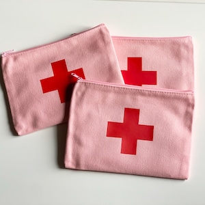 Puede incluir: Tres estuches rosas de lona con cremallera y un dise&ntilde;o de cruz roja grande. Los estuches est&aacute;n colocados sobre una superficie blanca. Las cremalleras son rosas.