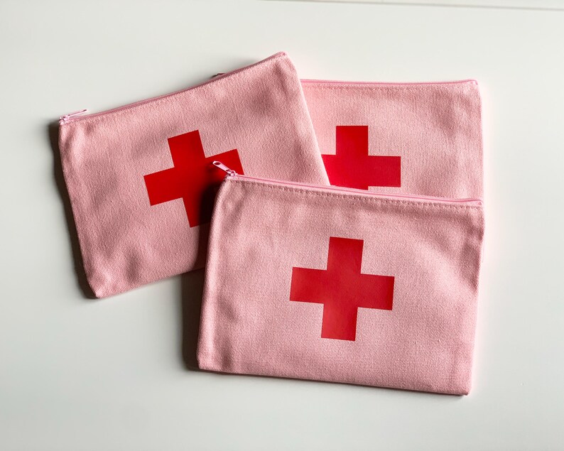 Puede incluir: Tres estuches rosas de lona con cremallera y un dise&ntilde;o de cruz roja grande. Los estuches est&aacute;n colocados sobre una superficie blanca. Las cremalleras son rosas.