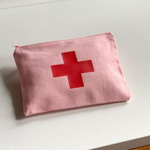 Puede incluir: Un estuche de lona rosa con una cruz roja. La bolsa rectangular tiene cierre de cremallera y est&aacute; dise&ntilde;ada para guardar suministros de primeros auxilios. La cruz est&aacute; centrada en la parte delantera.