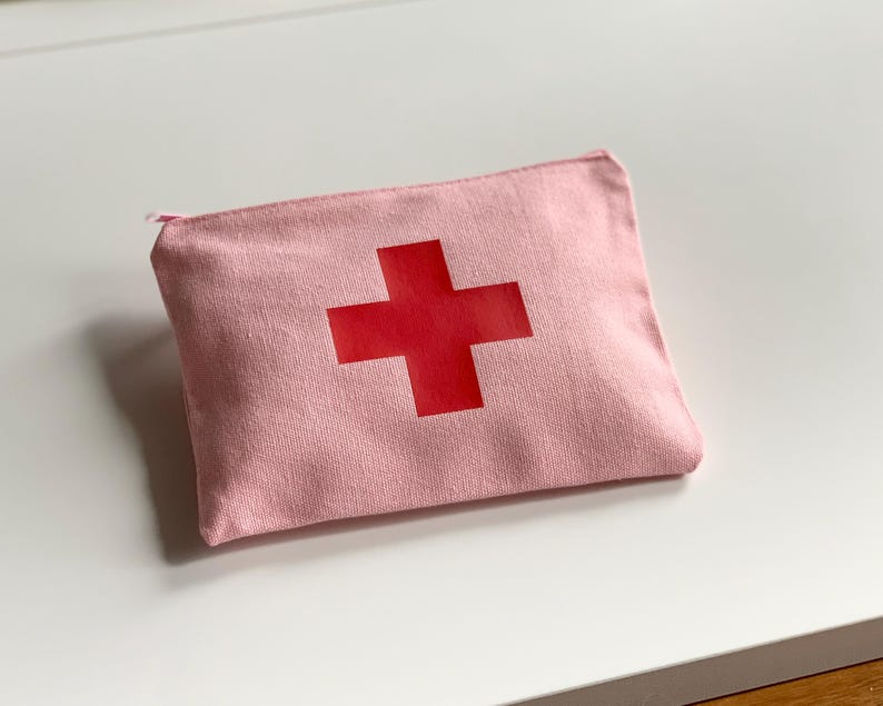 Puede incluir: Un estuche de lona rosa con una cruz roja. La bolsa rectangular tiene cierre de cremallera y est&aacute; dise&ntilde;ada para guardar suministros de primeros auxilios. La cruz est&aacute; centrada en la parte delantera.