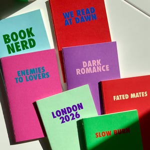 Peut inclure: Plusieurs cahiers colorés avec des textes audacieux. Les cahiers sont de différentes couleurs, notamment bleu, rouge, rose, violet et vert, et comportent des phrases telles que "BOOK NERD", "WE READ AT DAWN" et "ENEMIES TO LOVERS".