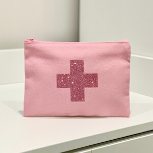 Puede incluir: Una bolsa de lona rosa con cremallera y un dise&ntilde;o de cruz brillante en el centro. La bolsa rectangular est&aacute; sobre una superficie blanca. La cruz es de un tono rosa m&aacute;s oscuro que la bolsa.