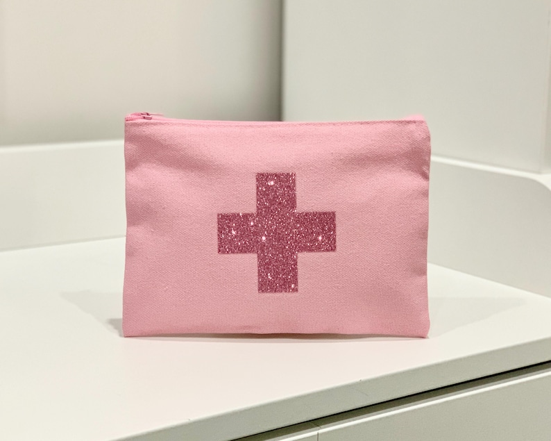 Puede incluir: Una bolsa de lona rosa con cremallera y un dise&ntilde;o de cruz brillante en el centro. La bolsa rectangular est&aacute; sobre una superficie blanca. La cruz es de un tono rosa m&aacute;s oscuro que la bolsa.