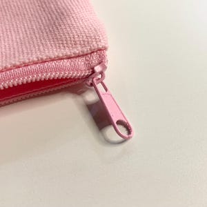 Puede incluir: Primer plano de una bolsa rosa con cremallera. La bolsa est&aacute; hecha de una tela texturizada y presenta una cremallera rosa a juego. El tirador de la cremallera tambi&eacute;n es rosa y tiene forma redondeada.
